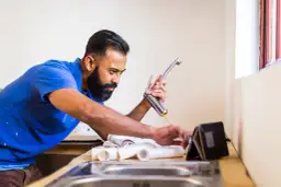 Hydraulik w niebieskiej koszulce montuje kran, patrząc na tablet.