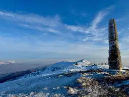 Kamienny obelisk na szczycie ośnieżonych **Rawki Bieszczady** pod błękitnym niebem.