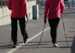 Dwie osoby w czerwonych kurtkach i czarnych spodniach idą z kijkami do nordic walking. Pokazują, jak ustawić kijki do chodzenia.