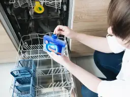 Osoba trzymająca w otwartej zmywarce niebieską butelkę z czyścikiem Finish.