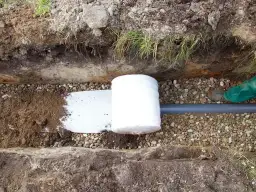 Budowa drenażu: rura drenarska owinięta geowłókniną, otoczona żwirem i ziemią, gotowa do zasypania.