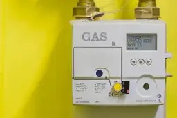Medidor de gas blanco con pantalla digital y conexiones doradas sobre fondo amarillo.