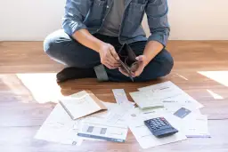 Mężczyzna siedzi na podłodze, przeglądając pusty portfel wśród rachunków, kalendarza i kalkulatora.