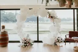 Okrągła girlanda z białych balonów ozdobiona kwiatami, obok beczki z tortem.