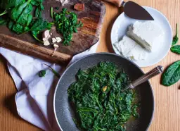 Jak doprawić szpinak? Na desce świeży szpinak, czosnek i ser feta na talerzu. W patelni gotowy, duszony szpinak.