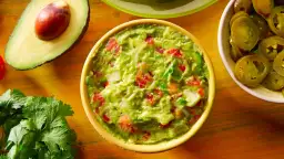 Miseczka guacamole z awokado, pomidorami i cebulą, obok połówka awokado i plasterki jalapeño.