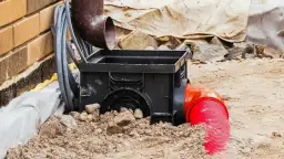 Czarny osadnik rynnowy w kostce, podłączony do czerwonej rury odpływowej, częściowo zasypany ziemią i kamieniami.