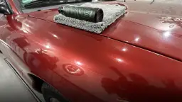 Butelka wosku samochodowego SWAG na szarej mikrofibrze leżącej na błyszczącym, czerwonym lakierze auta.
