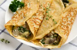 Naleśniki z mięsem i grzybami, z dodatkiem szpinaku, podane na białym talerzu z gałązkami tymianku i natki pietruszki.