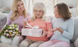 Trzy kobiety na kanapie: starsza otwiera prezent, młodsza trzyma bukiet, najmłodsza gestykuluje.