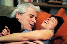 Ein Mann mit grauem Haar und eine lachende Frau liegen eng beieinander auf einem orangefarbenen Kissen.