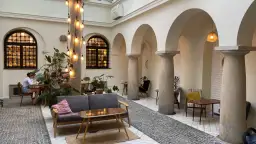 Przytulna kawiarnia patio z łukami i kolumnami. Goście siedzą przy stolikach, a obok stoi wygodna sofa. Widać rośliny i dekoracyjne lampki. Menu czeka na odkrycie.