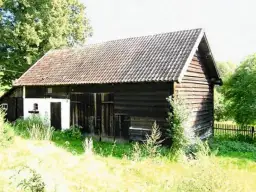 Drewniany budynek gospodarczy z ciemną elewacją i dachówką, otoczony zielenią.