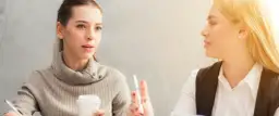 Dwie kobiety rozmawiają przy stole, jedna trzyma kubek kawy.