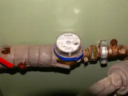 Ktoś naprawia zawór przed wodomierzem. Widoczny jest zardzewiały metalowy element i izolacja rury.