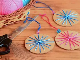 Kolorowe bombki z papieru dla dzieci, ozdobione nitkami, gotowe do zawieszenia. Obok nożyczki i koszyk z włóczką.
