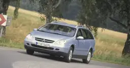 Jasnoniebieskie kombi Citroen na zakręcie drogi, z drzewami i polem w tle.