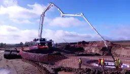 Ogromna pompa do betonu pracuje na budowie, dostarczając materiał do wykopu. Pracownicy w kamizelkach obserwują proces.