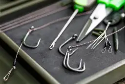 Zbliżenie na haczyki karpiowe, żyłkę, nożyczki i inne akcesoria wędkarskie w pudełku.