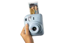 Niebieski aparat Instax Mini 12, trzymany w dłoni, z właśnie wysuniętym, jasnym zdjęciem dwóch uśmiechniętych przyjaciółek.