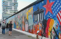 Bunte East Side Gallery in Berlin mit Graffiti und Menschen, im Hintergrund ein modernes Hochhaus.