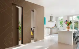 Nowoczesne drzwi ukryte w ścianie, z subtelnym wzorem i listwą przypodłogową, prowadzą do jasnej kuchni z wyspą i bukietem kwiatów.