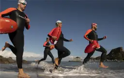 Trzy osoby w piankach biegają po plaży z pomarańczowymi bojami.