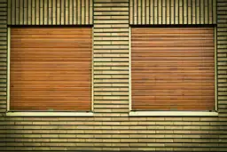 Dwie brązowe rolety okienne na żółtej ceglanej ścianie. Dowiedz się, jak naprawić roletę, gdy się zablokuje.