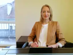 Kobieta w beżowej marynarce siedzi przy biurku i pisze.