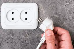 Dłoń trzyma wtyczkę do podłączenia pralki. Gniazdko elektryczne znajduje się na ścianie.