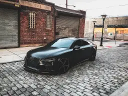 Matowy czarny Audi RS7 na brukowanej ulicy przed ceglanym budynkiem.