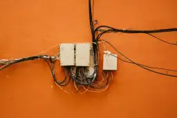 Stare skrzynki elektryczne z plątaniną kabli na pomarańczowej ścianie.