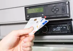 Dłoń wkłada kartę kierowcy do tachografu. Widoczny slot na kartę i ekran urządzenia.
