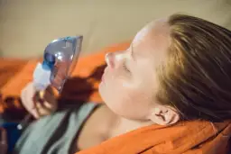 Kobieta z maską inhalatora na twarzy, leżąca na pomarańczowym tle.