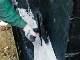 Ręka w rękawicy nakłada białą masę na czarną, wilgotną ścianę. To szlam mineralny, który chroni przed wodą.