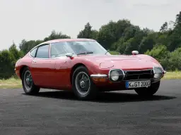 Klasyczny, czerwony Toyota 2000GT, jeden z najbardziej pożądanych japońskich samochodów sportowych, prezentuje swoje eleganckie linie na tle zieleni.