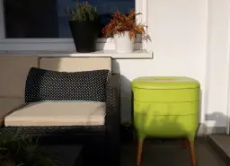 Zielony kompostownik na balkonie, obok wygodnej kanapy z poduszką w grochy.