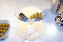 Kto wybierze Dirty Martini czy klasyczne Dry Martini? Kieliszek z oliwkami i drinkiem na białym tle.