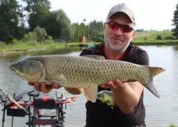 Wędkarz w czapce i okularach pokazuje dużą rybę na tle jeziora i sprzętu wędkarskiego.