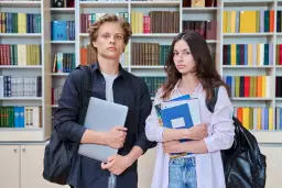 Dwóch studentów z książkami i laptopem na tle regałów z książkami.