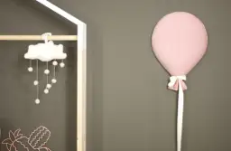 Różowy balon z kokardką przyczepiony do szarej ściany obok dziecięcej dekoracji.