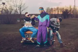 Trzech mężczyzn w strojach z lat 90. moda, siedzących na starym samochodzie. Jeden w dresie, drugi w bluzie i jeansach.