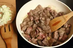 Smażona wątróbka w patelni z drewnianą łopatką, obok posiekana cebula.