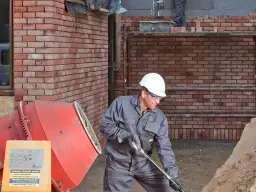 Budowlaniec w kasku pracuje przy betoniarce. Obok stos piasku.