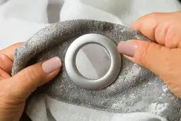 Dłonie z różowym lakierem do paznokci trzymają szarą tkaninę z wzorem, pokazując srebrne kółka do firan.