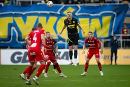 Piłkarz w żółto-czarnej koszulce wyskakuje do piłki, podczas gdy zawodnicy w czerwonych strojach obserwują. Czy to oni są tymi, kto spada z ekstraklasy?