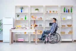 Mężczyzna na wózku inwalidzkim w kuchni, która jest przystosowana jako mieszkanie dla niepełnosprawnych. Wygodny dostęp do półek i stołu.