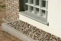 Kamienna opaska wokół domu inspiracje: żwirowa rabata z kamieni polnych przy betonowym murku i ceglanej ścianie.