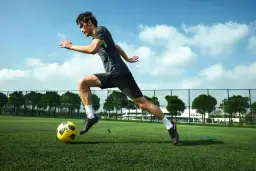 Piłkarz w ruchu kopie żółtą piłkę na zielonym boisku pod błękitnym niebem.