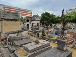 Kamienne nagrobki i mauzolea na cmentarzu w Paryżu, z nowoczesnym budynkiem w tle.